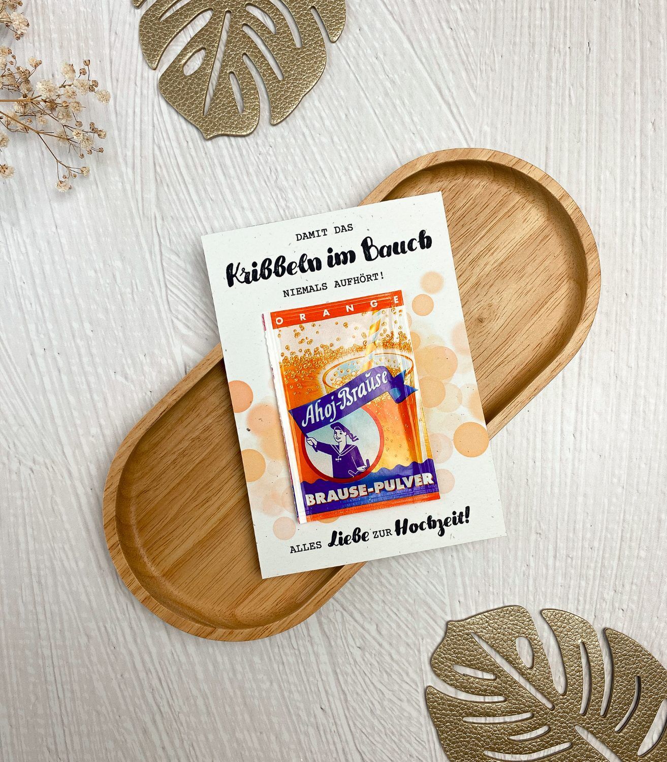 Eine ausgedruckte Karte mit einer aufgeklebten Ahoj Brause mit Geschmack Orange, im Hintergrund sind orangefarbene Blubberblasen. Auf der Karte steht der Spruch "Damit das Kribbeln im Bauch niemals aufhört. Alles Liebe zur Hochzeit".