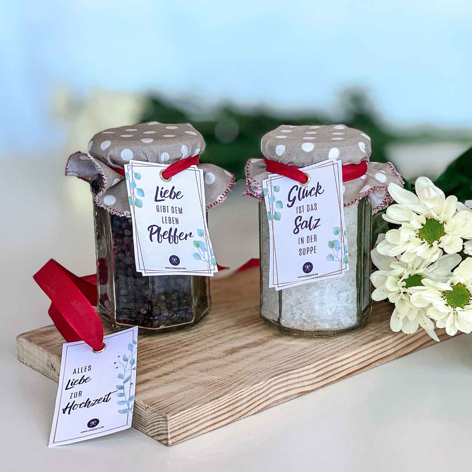 Fertiges Geschenkset Salz und Pfeffer zur Hochzeit Zwei dekorierte Gläser mit Salz und Pfeffer zur Hochzeit und roten Schleifen sowie einem zusätzlichen Anhänger Alles Liebe zur Hochzeit auf einem Holzbrett.