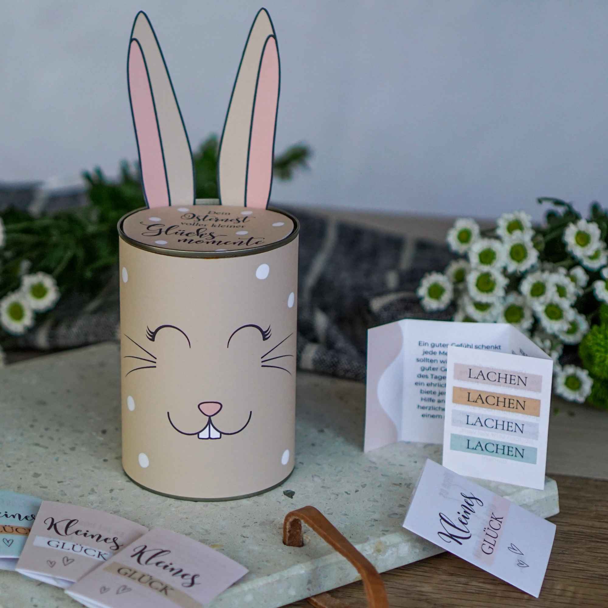 Eine beige Konservendose im Hasen-Look mit der Aufschrift Osternest voller kleiner Glücksmomente und einem geöffneten Kärtchen mit dem Wort Lachen als kreatives Ostergeschenk.