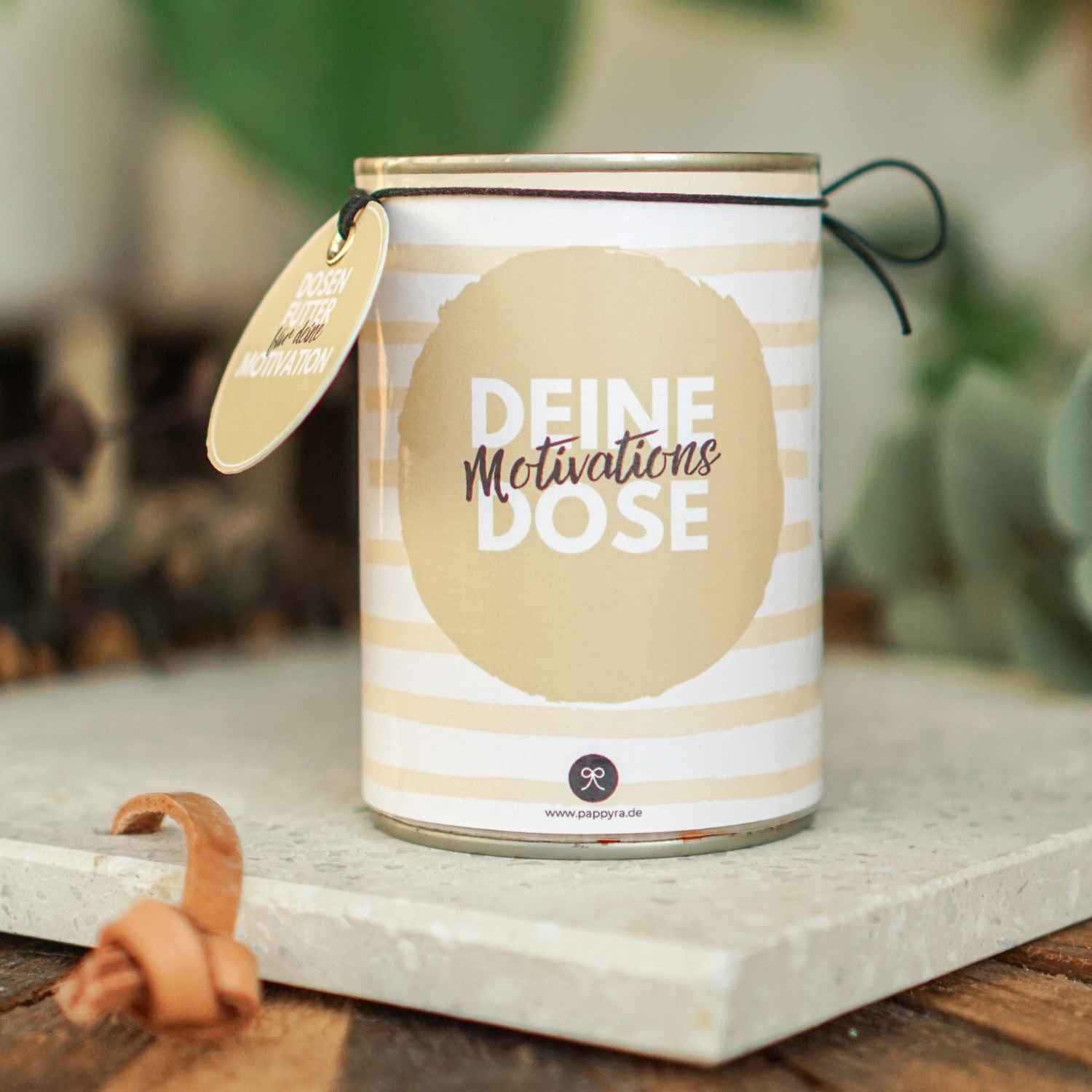 Eine fertig gebastelte Konservendose mit beiger Streifen-Banderole und Anhänger, die als kreatives Motivation Geschenk auf einem Steinsockel steht.