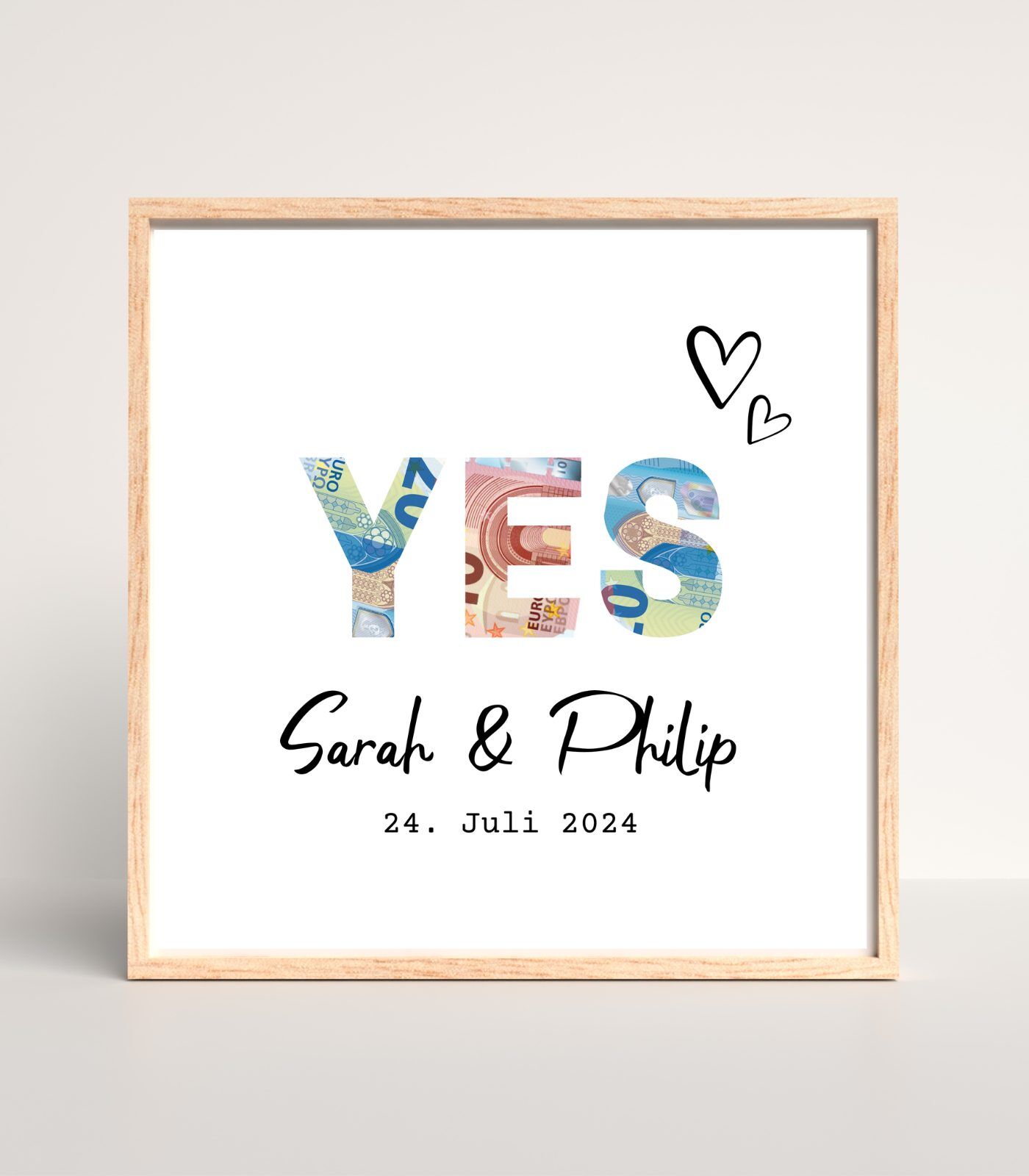 Quadratisches Poster mit den Namen des Brautpaars und dem Hochzeitsdatum. Oberhalb stehen die Buchstaben "YES". Die Buchstaben wurden ausgeschnitten und hinter ihnen scheinen Geldscheine hindurch.